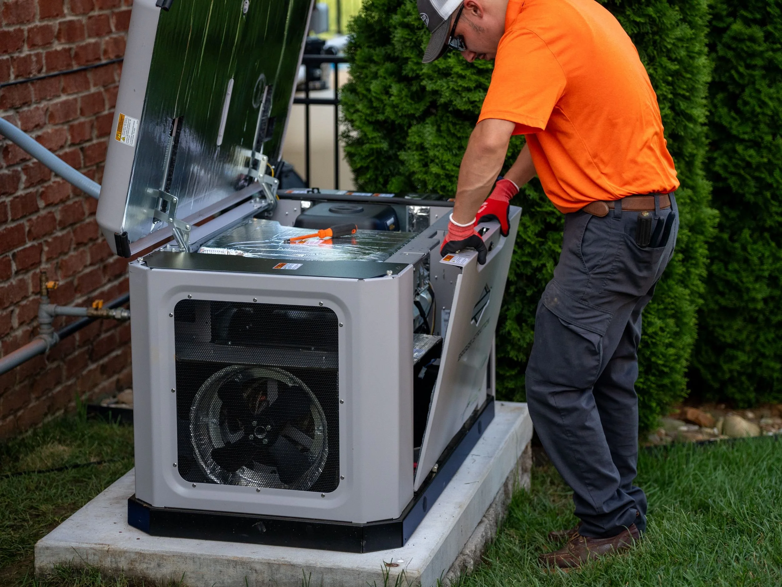A man in orange shirt and dark pants working on solar panel installation outside next to a brick wall and green bush. He is wearing gloves, a baseball cap, and sunglasses.