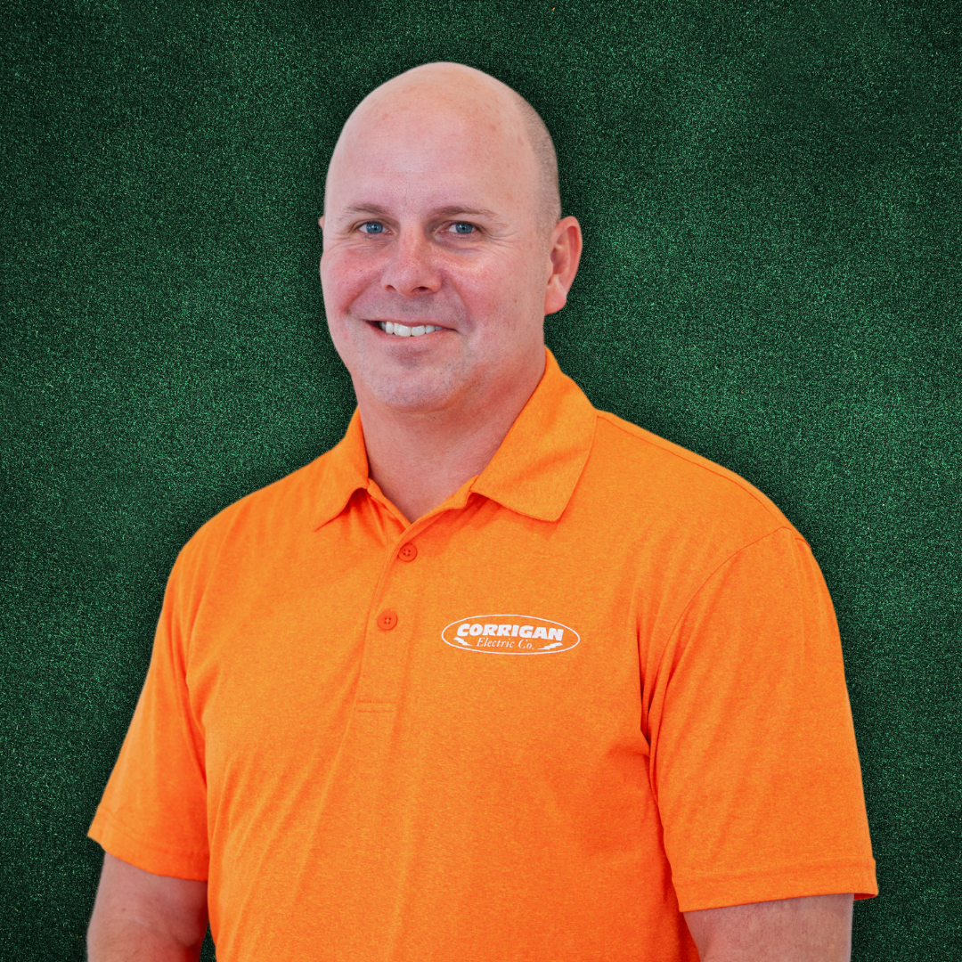 A smiling man with a shaved head wearing an orange polo shirt with a logo that says "Corrigan Electric Co." against a green background.