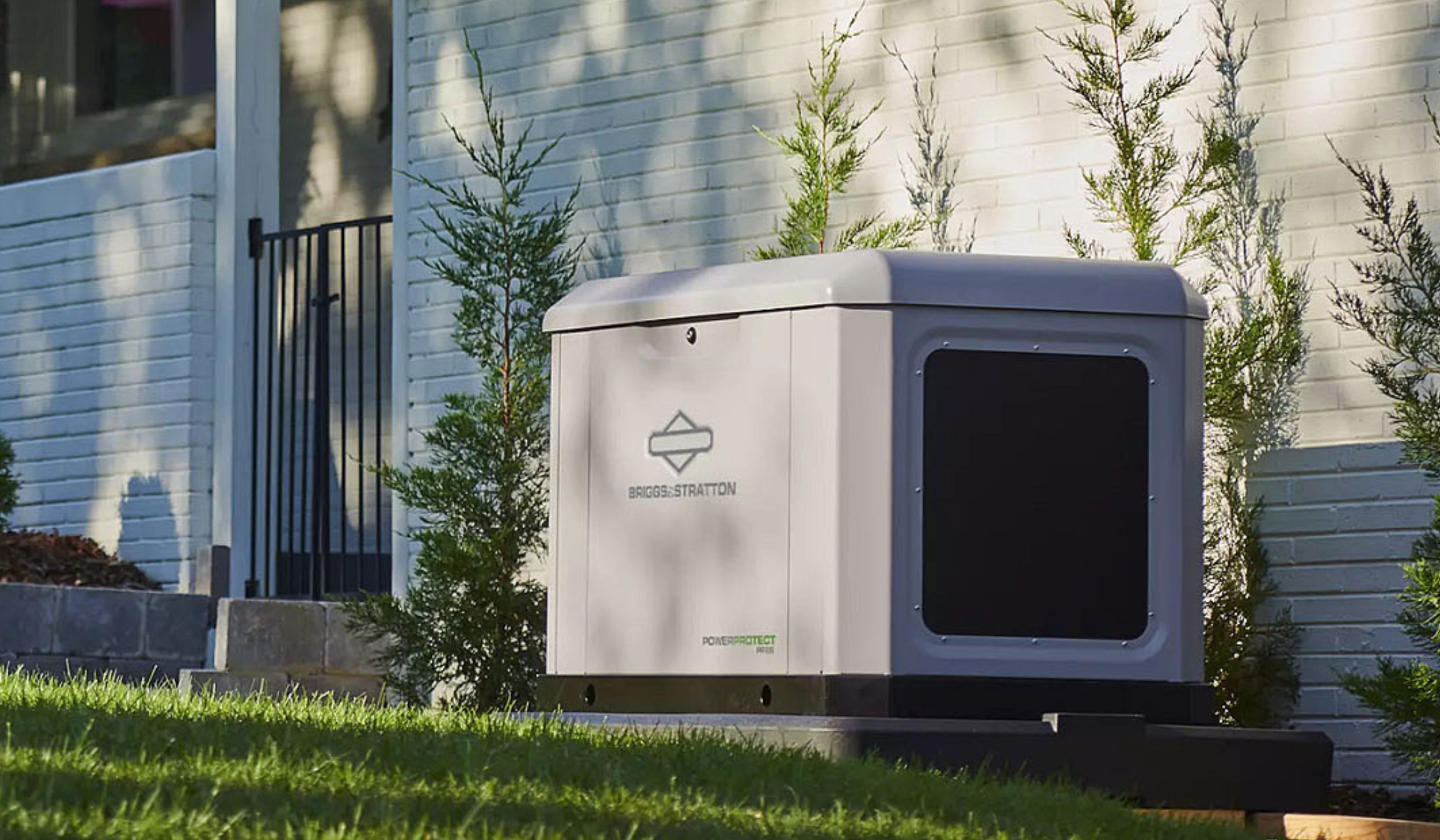 An outdoor scene showing a white brick house wall with a black gate, small trees, and a gray outdoor electrical box labeled "Briggs & Stratton" and "PowerProtect".