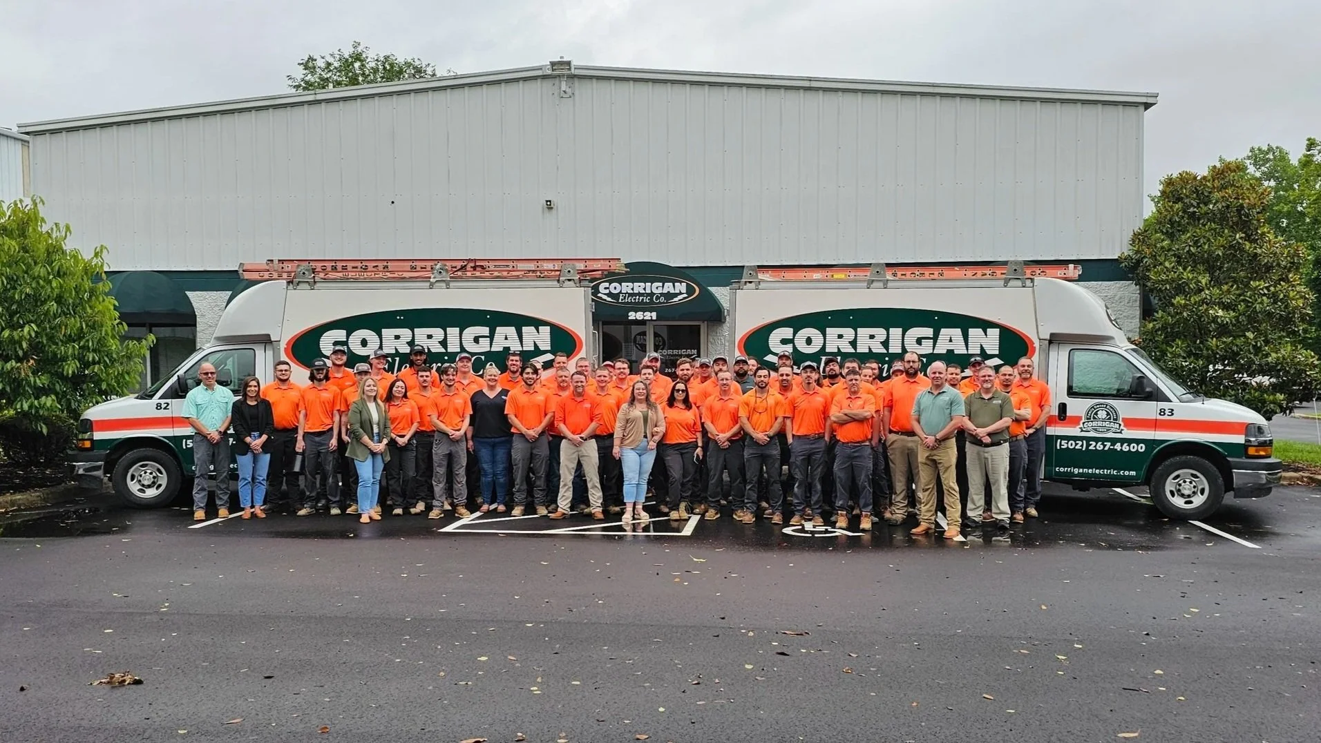 A large group of people standing in front of company trucks. The people are wearing orange shirts, and the trucks have the logo 'Corrigan Electric' on them.