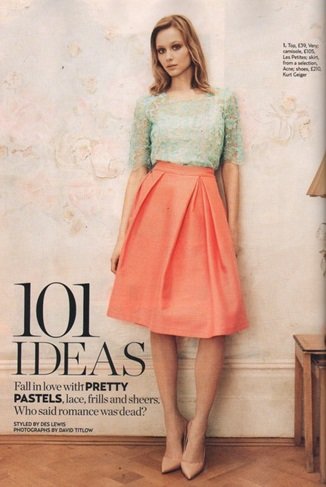 A woman standing against a light-colored wall in front of a magazine cover, wearing a pastel green top, peach-colored skirt, and beige high heels.