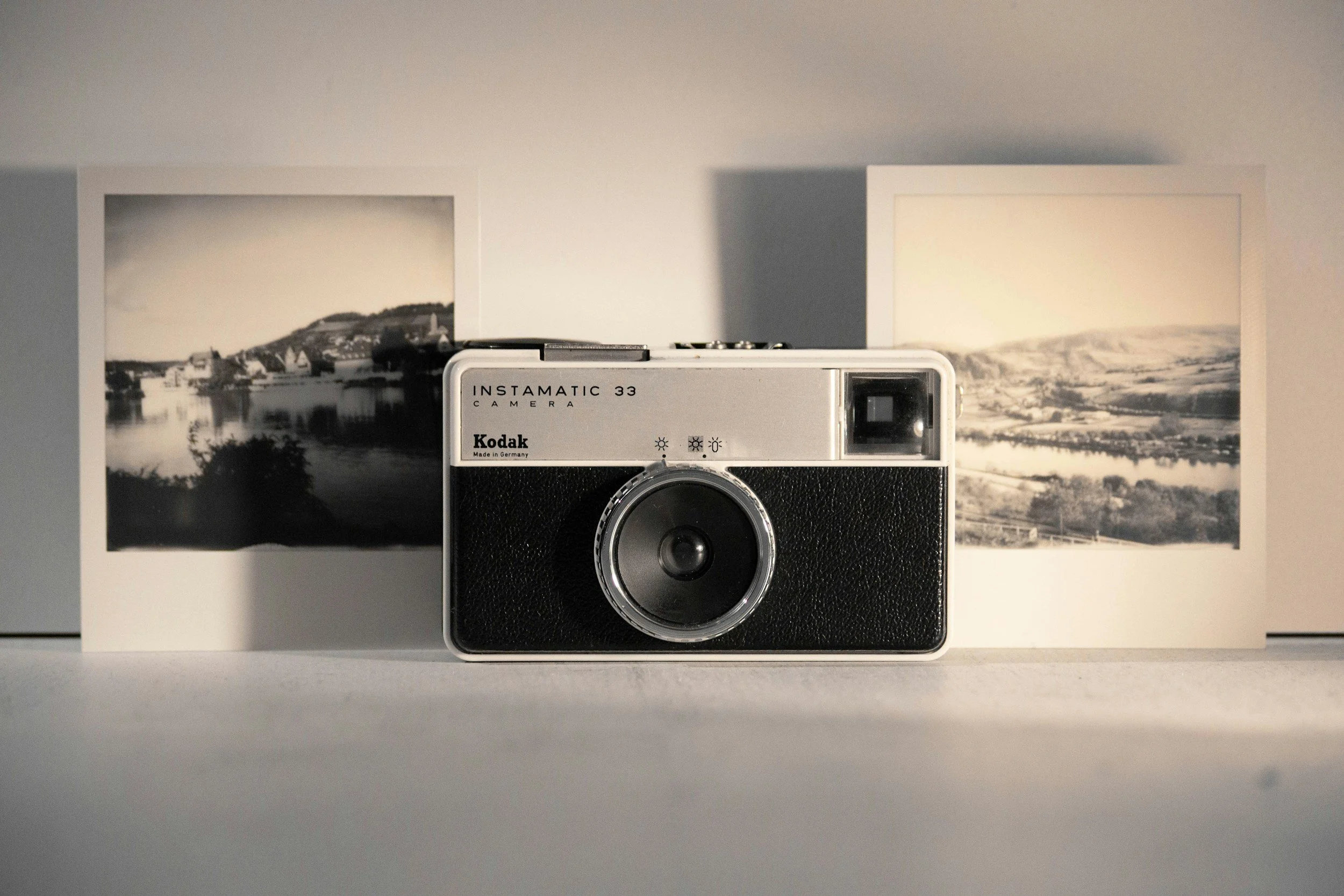 A vintage Kodak Instamatic 33 camera placed on a white surface, with black and white landscape photographs in the background.