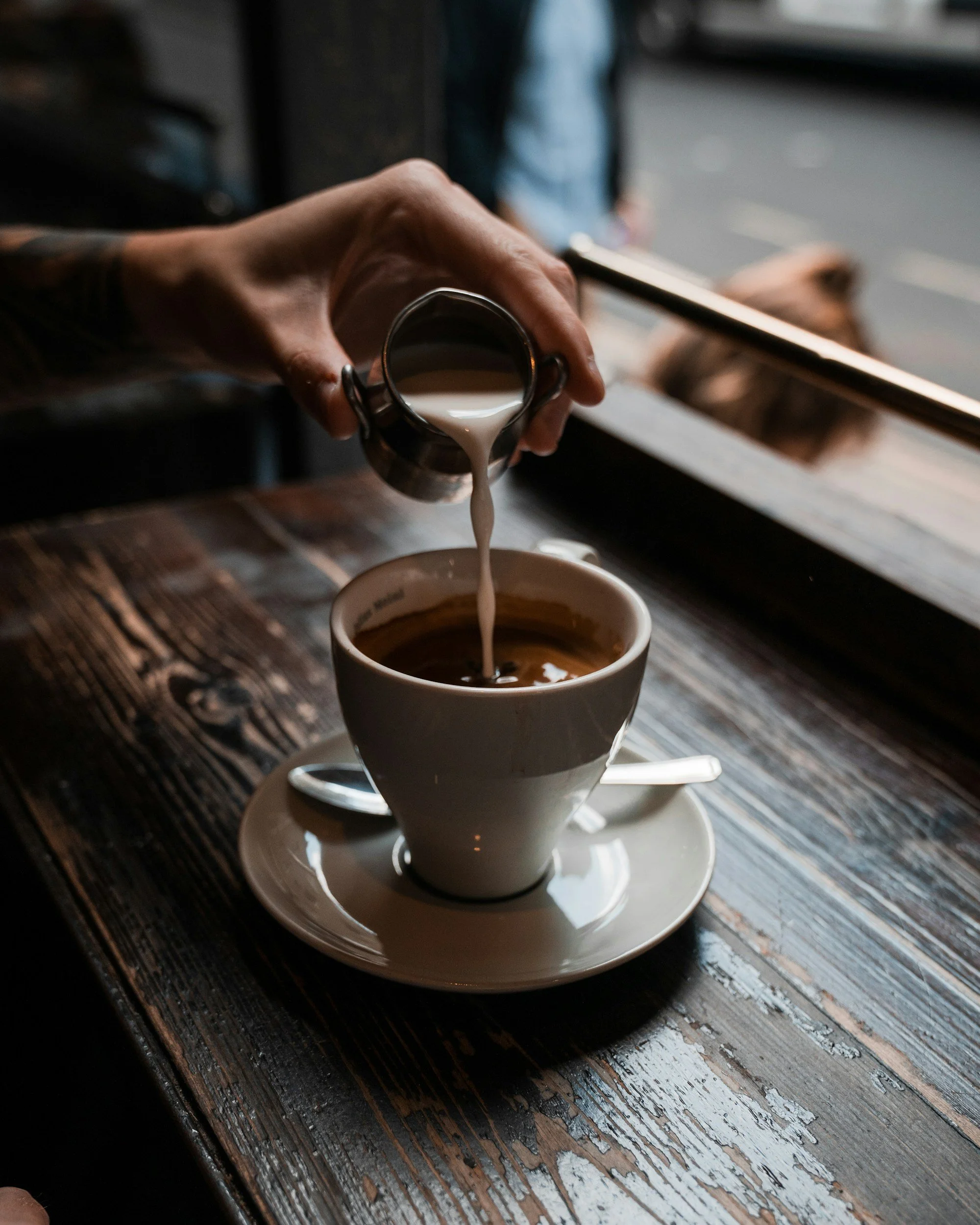 A person is pouring cream into a cup of coffee on a wooden table at a café.