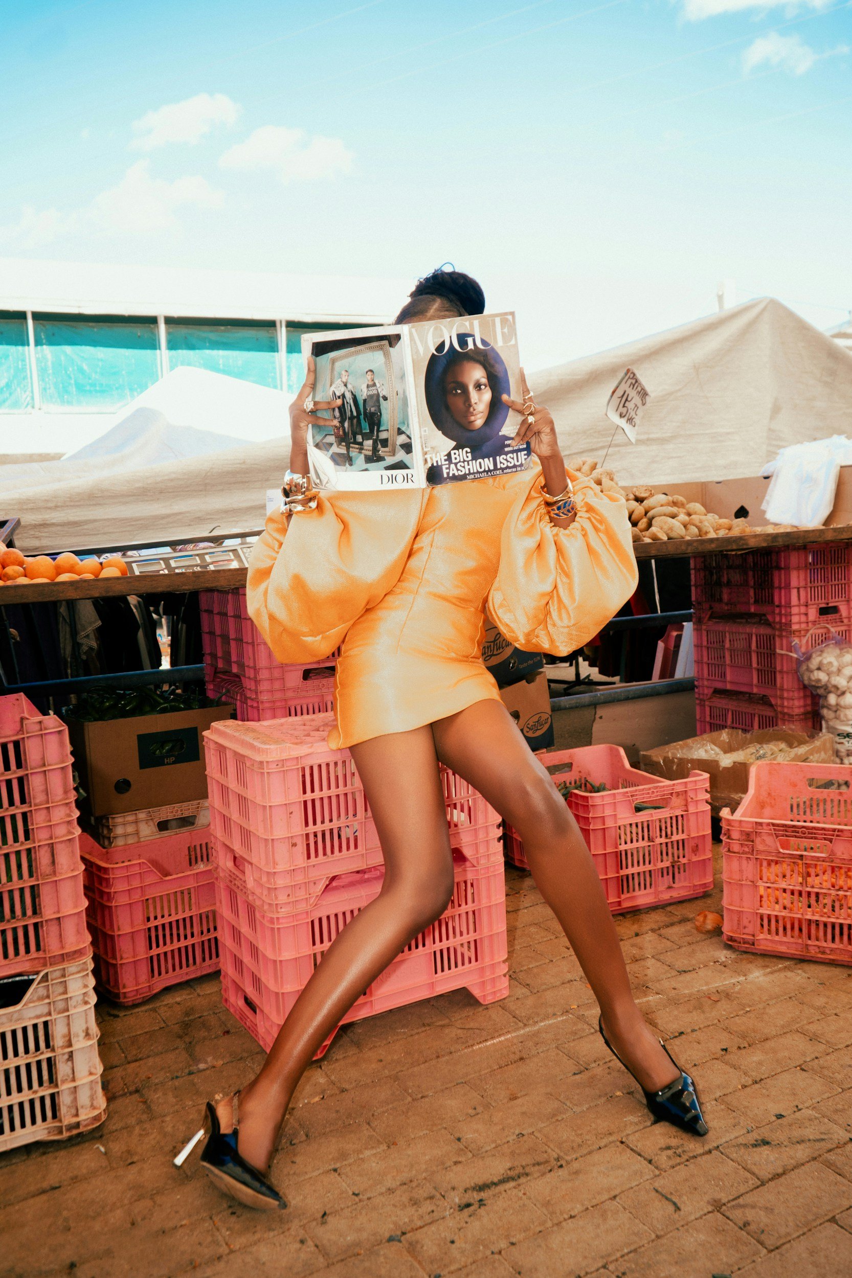 A person with tanned legs wearing high heels, a yellow dress, and a gold bracelet holding a Vogue magazine covering their face, standing amidst pink crates of produce at an outdoor market.