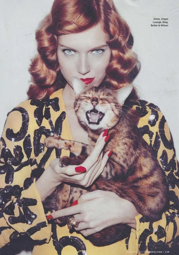 A woman with red curly hair and red lipstick holding a laughing tabby cat with orange and black stripes.