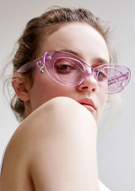 A woman with light skin, brown hair, and purple sunglasses looking over her shoulder, with a neutral background.