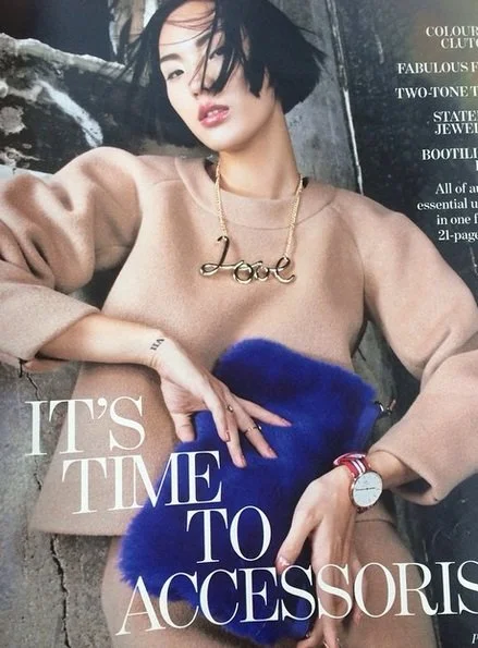 Close-up of a woman taking a selfie, wearing a beige sweater, a gold necklace spelling "Love," and holding a blue accessory. Magazine cover with the headline "It's time to accessorize."