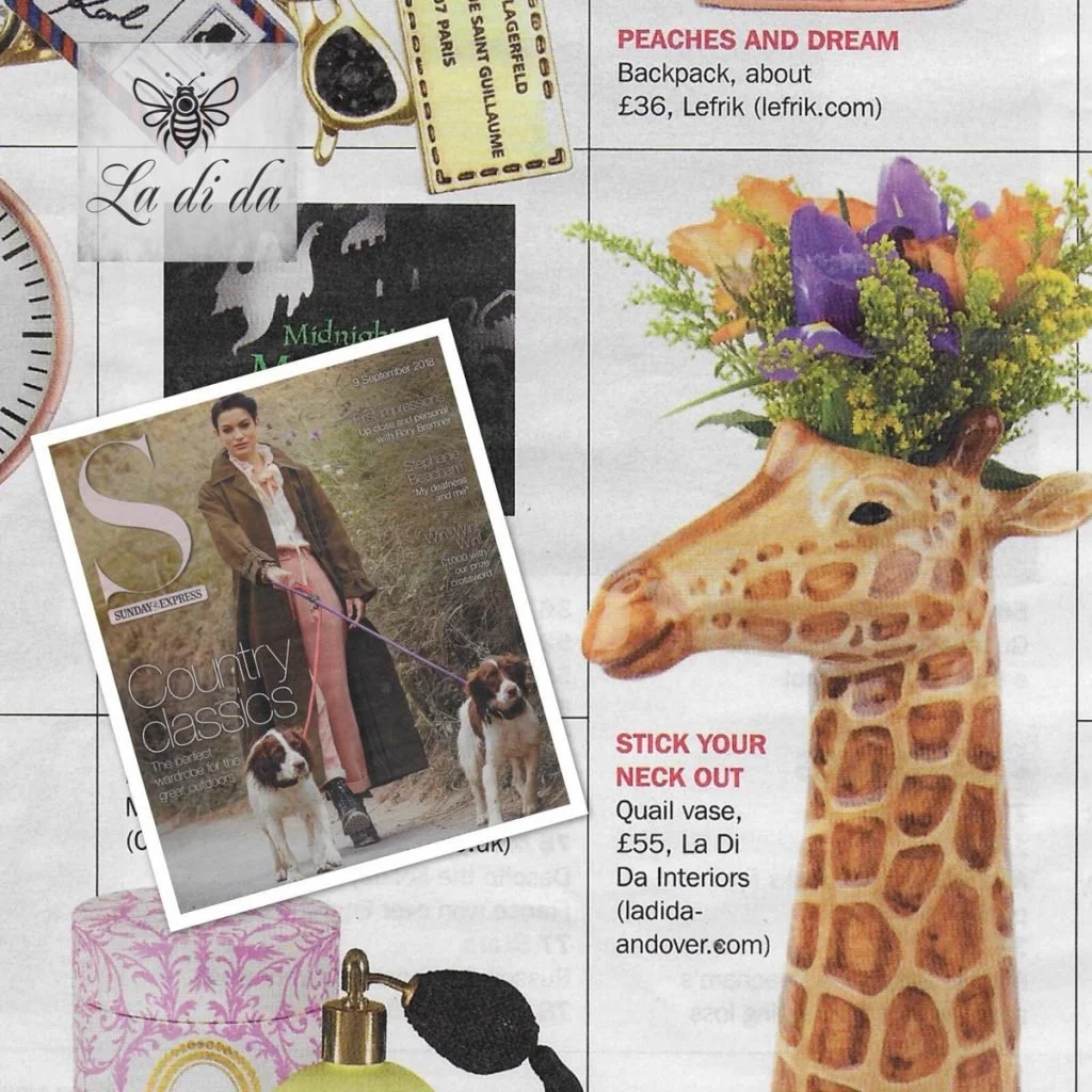 A magazine page featuring a giraffe-shaped vase filled with purple and orange flowers on a white background. The vase has a long neck and realistic giraffe features. Nearby, there is a small illustrated package with a bee logo, and part of a perfume 