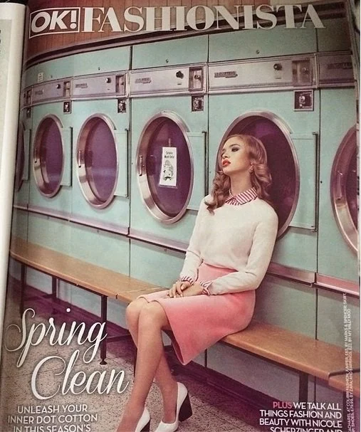 A woman sitting on a bench beside a row of washing machines in a laundromat, looking to her left. She has wavy brown hair, is wearing a white sweater with a pink and white striped collar, a pink skirt, and white heels. The magazine cover features the