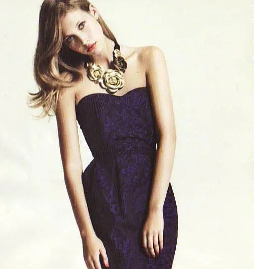 A young woman with wavy hair wearing a strapless purple dress and a statement necklace with large floral design.