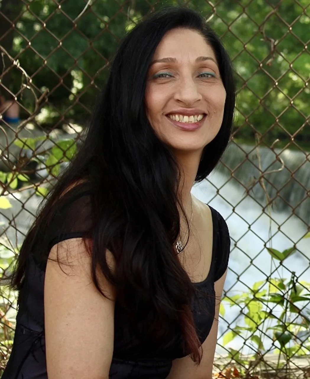 Smiling woman with long black hair standing outdoors near a chain-link fence with greenery and water in the background.