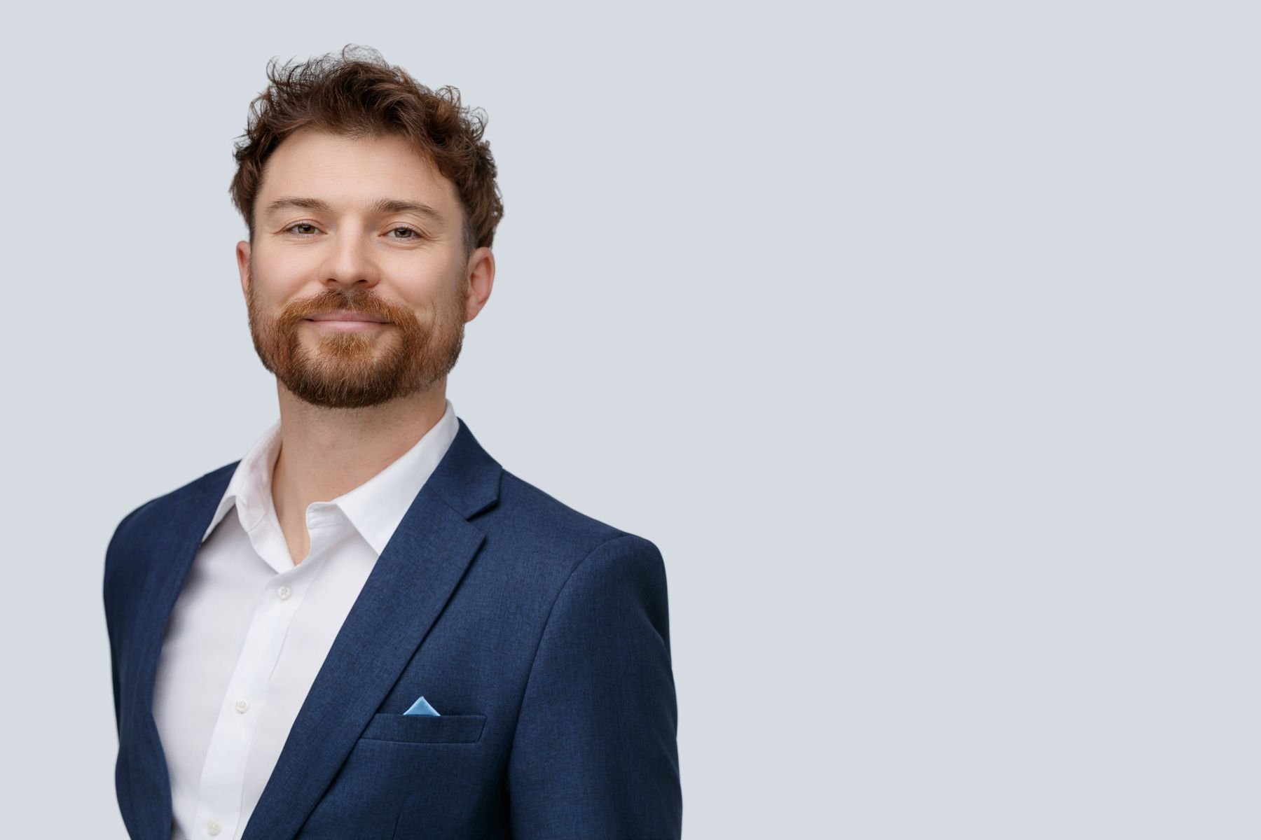 Portrait of a man with a beard and short, curly brown hair, dressed in a blue suit and white shirt, standing against a light gray background.