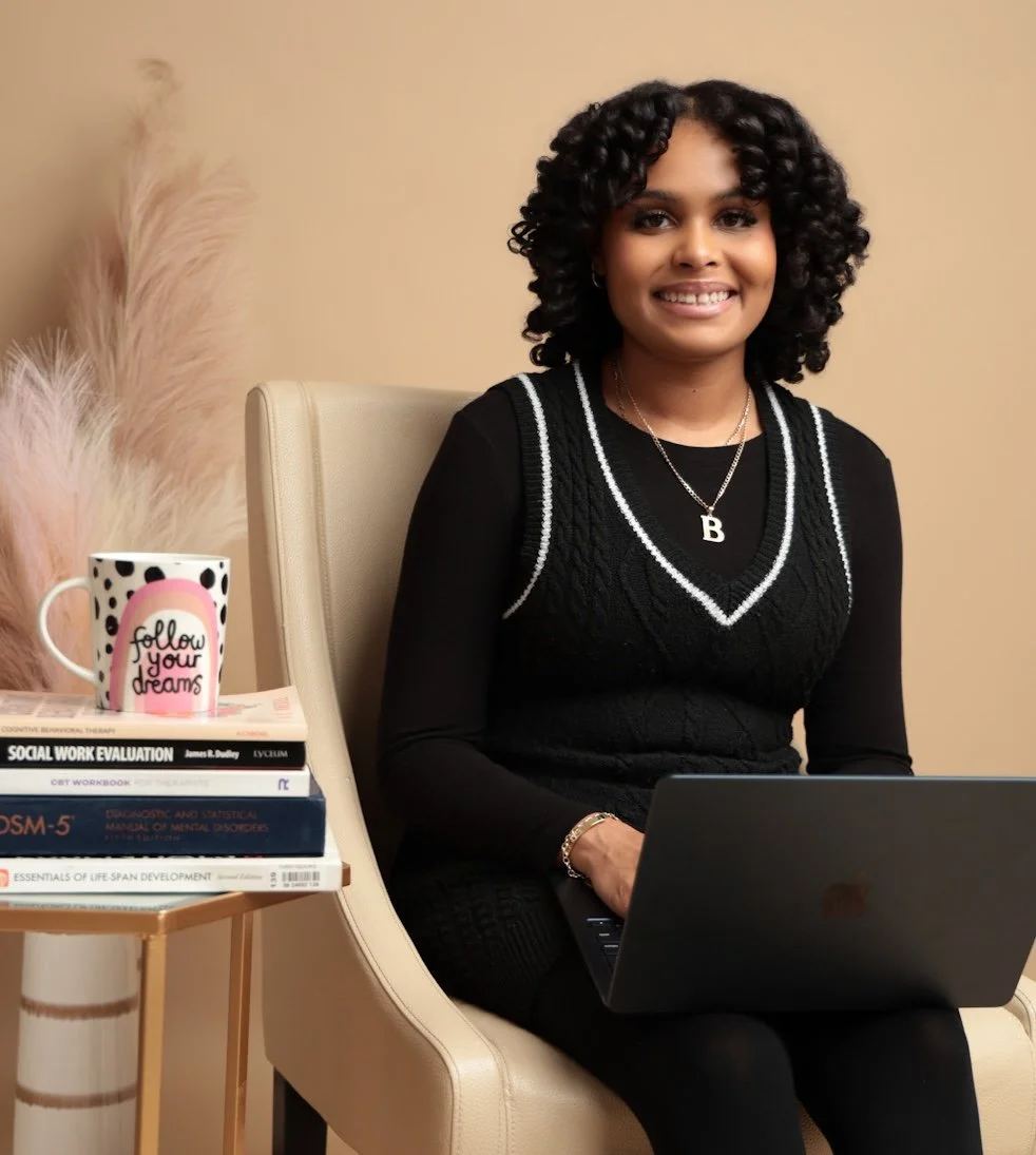 A young woman with curly black hair sitting on a beige chair with a laptop on her lap. Beside her is a small side table with books, a mug that says 'follow your dreams,' and a tall arrangement of pampas grass. She is wearing a black sweater vest with white trim, a necklace with the letter B, and smiling at the camera.