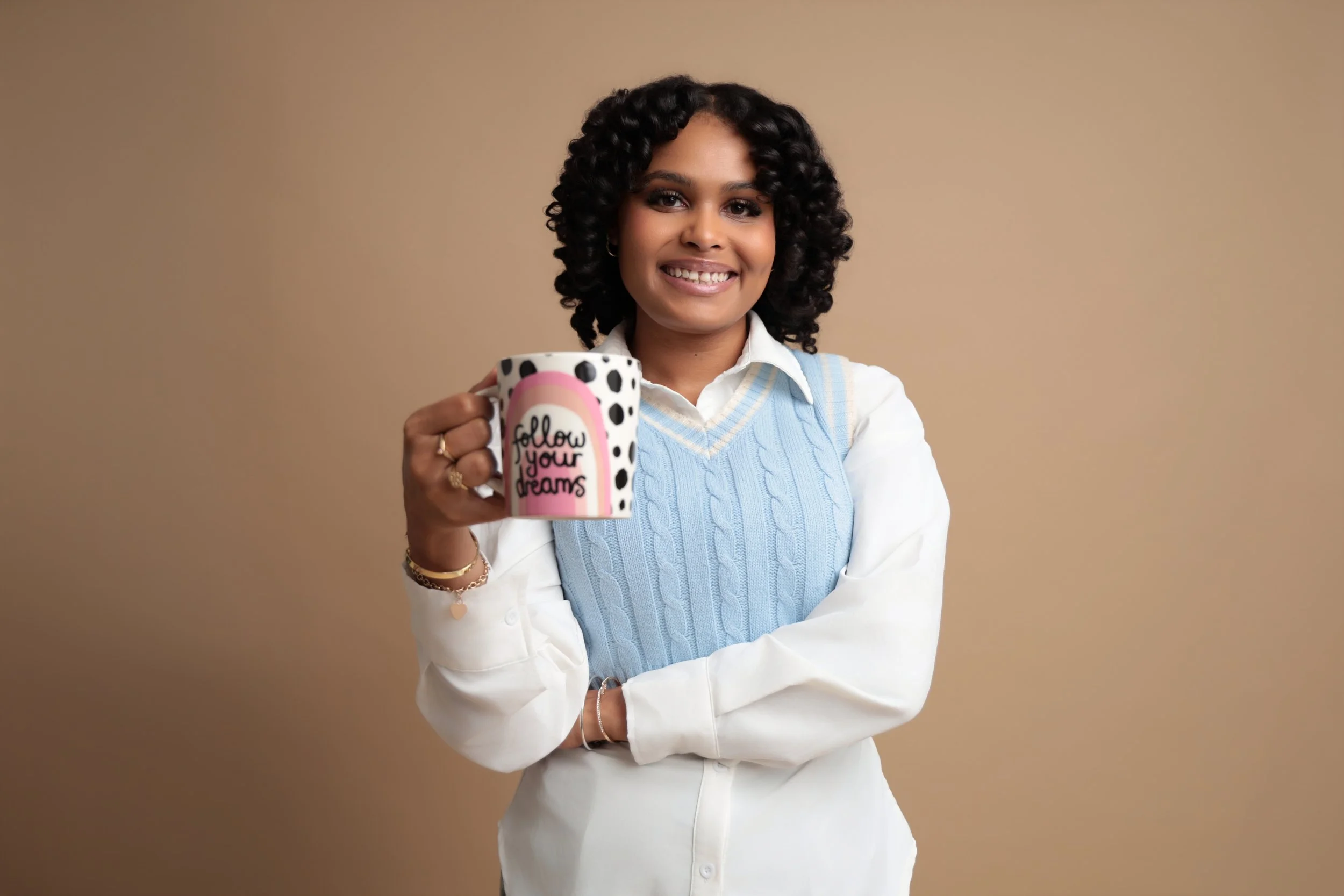 A smiling woman with curly black hair wearing a white shirt and a light blue sweater vest, holding a ceramic mug with polka dots and the words 'follow your dreams' written on it, standing against a beige background.