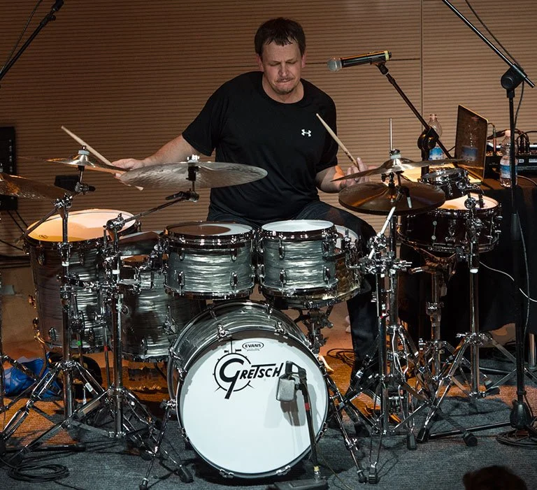 A man playing a Gretsch drum set in a studio with wooden panel walls, wearing a black Under Armour shirt, focused on playing.
