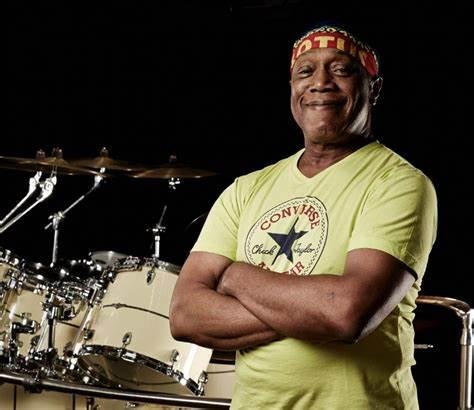 Smiling African American man wearing a colorful headband and a yellow t-shirt with a graphic, standing with crossed arms behind a drum set against a black background.