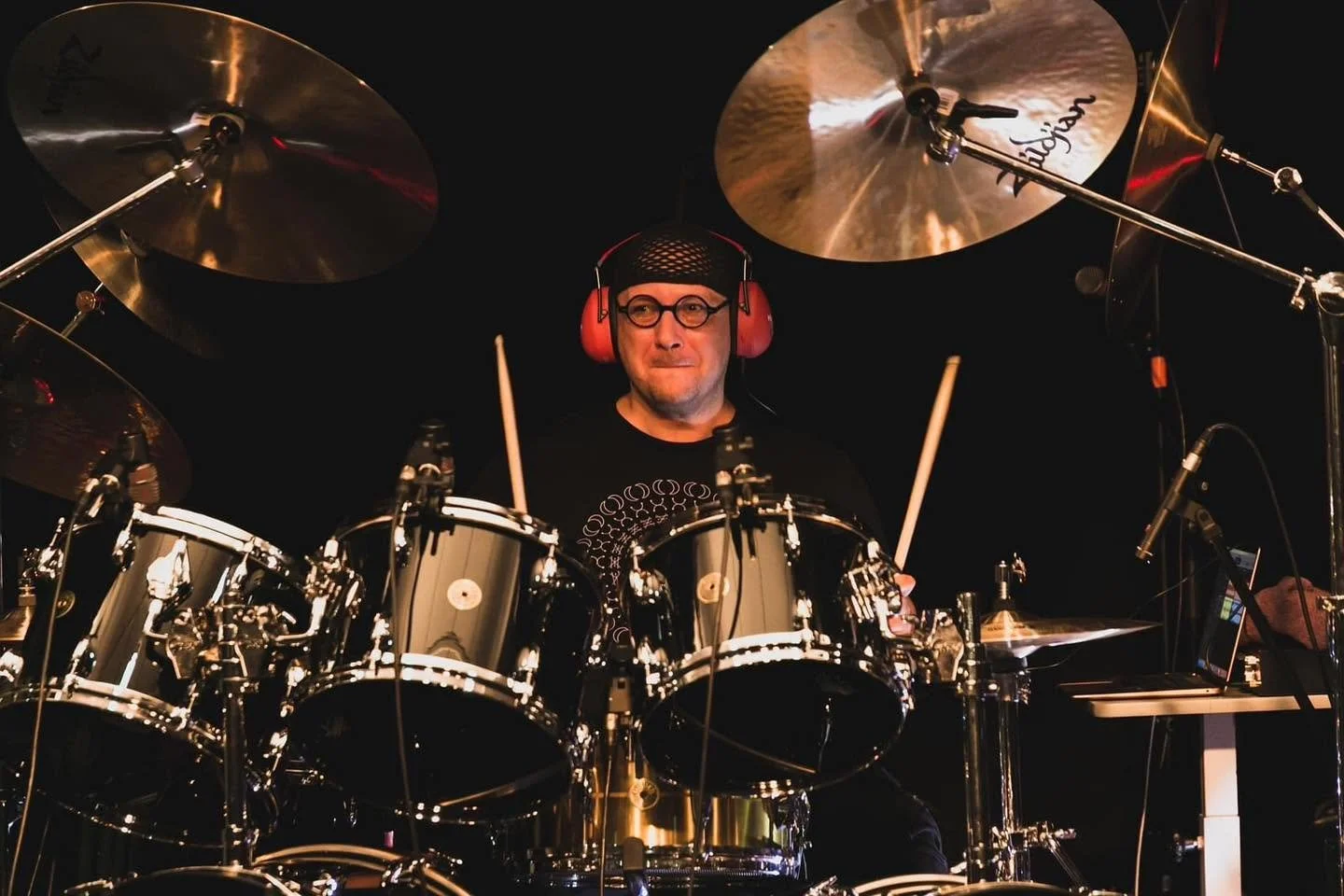 A man wearing a black cap and glasses playing a drum set, with red protective headphones, is illuminated against a dark background.