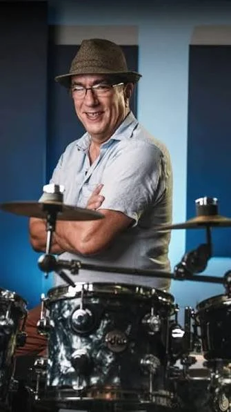 A man wearing glasses, a brown fedora, and a light-colored shirt, smiling and standing behind a drum set in a music studio.