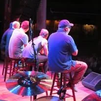 Four people sitting on bar stools on a stage, watching a performance in a dimly lit setting.