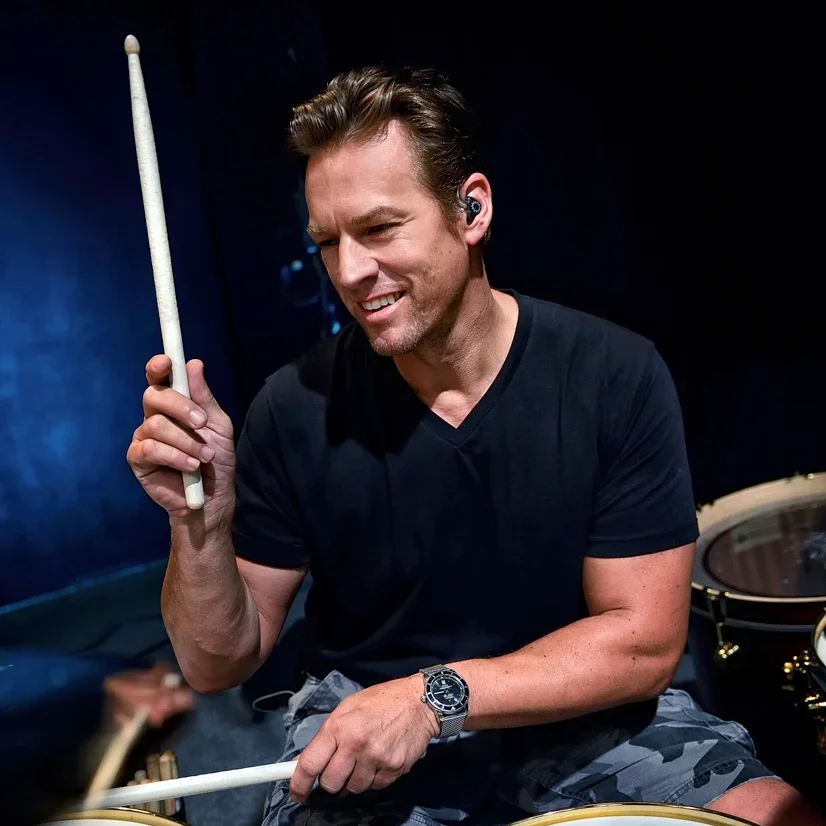 A man playing drums, holding drumsticks, smiling, wearing a black t-shirt and a watch, with a dark background.