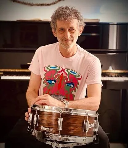 A man with curly gray hair and a beard sitting in front of a piano, holding a drum.