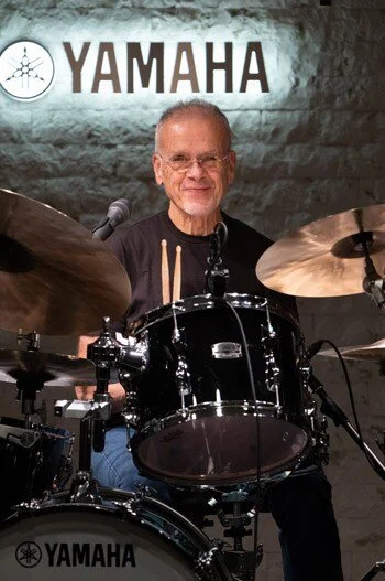 Older man with glasses sitting behind a black Yamaha drum set, smiling, with a Yamaha logo and a brick wall in the background.