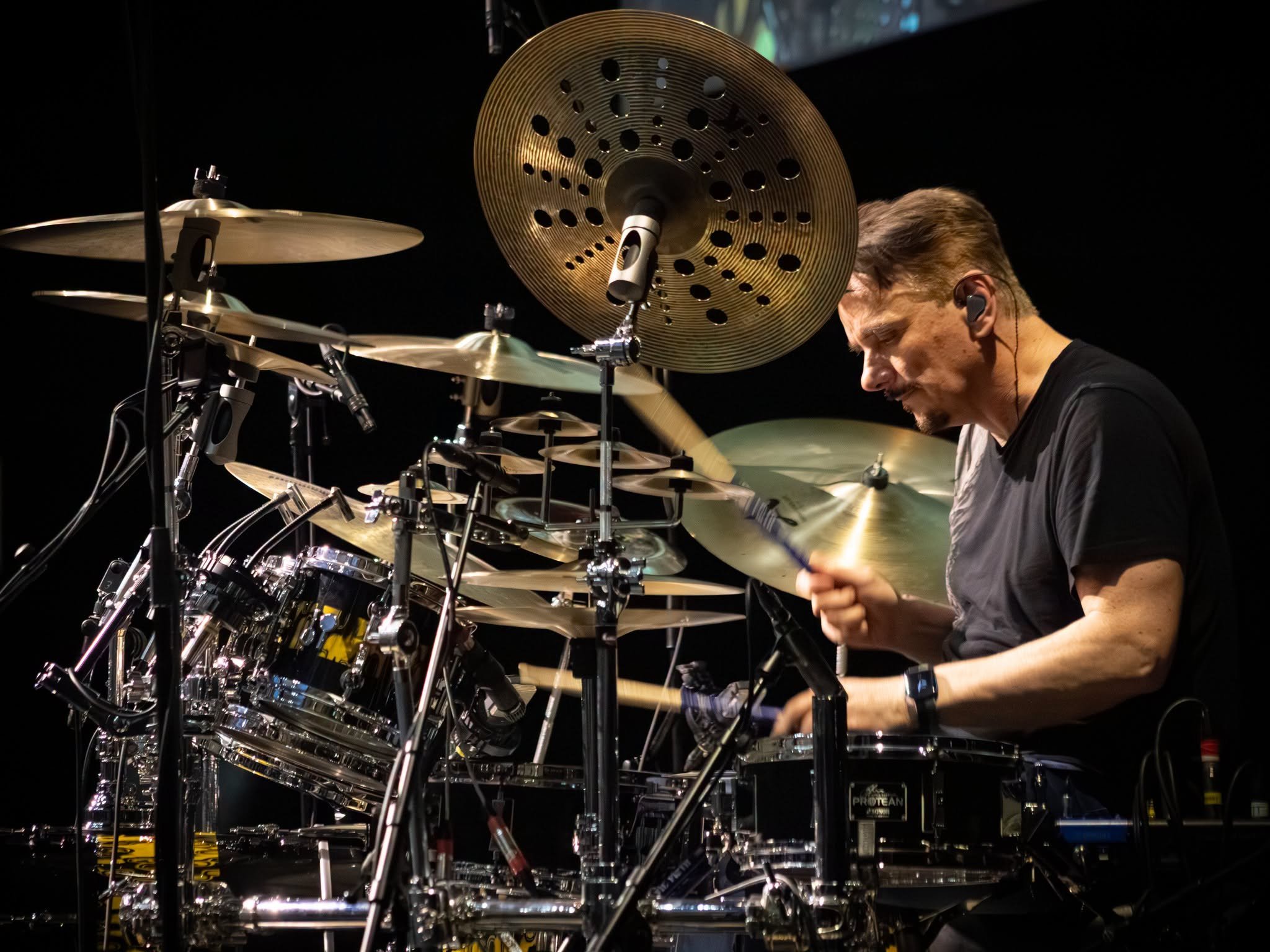 A drummer playing drums on stage with a dark background, focus on cymbals and drum kit, with a man wearing an in-ear monitor.