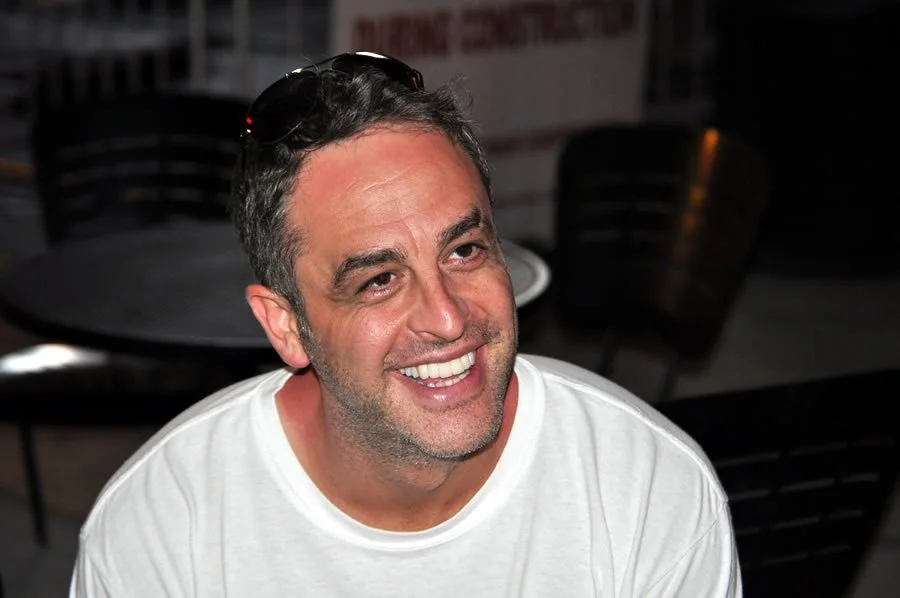 Smiling man with sunglasses on his head, wearing a white T-shirt, in a casual setting.