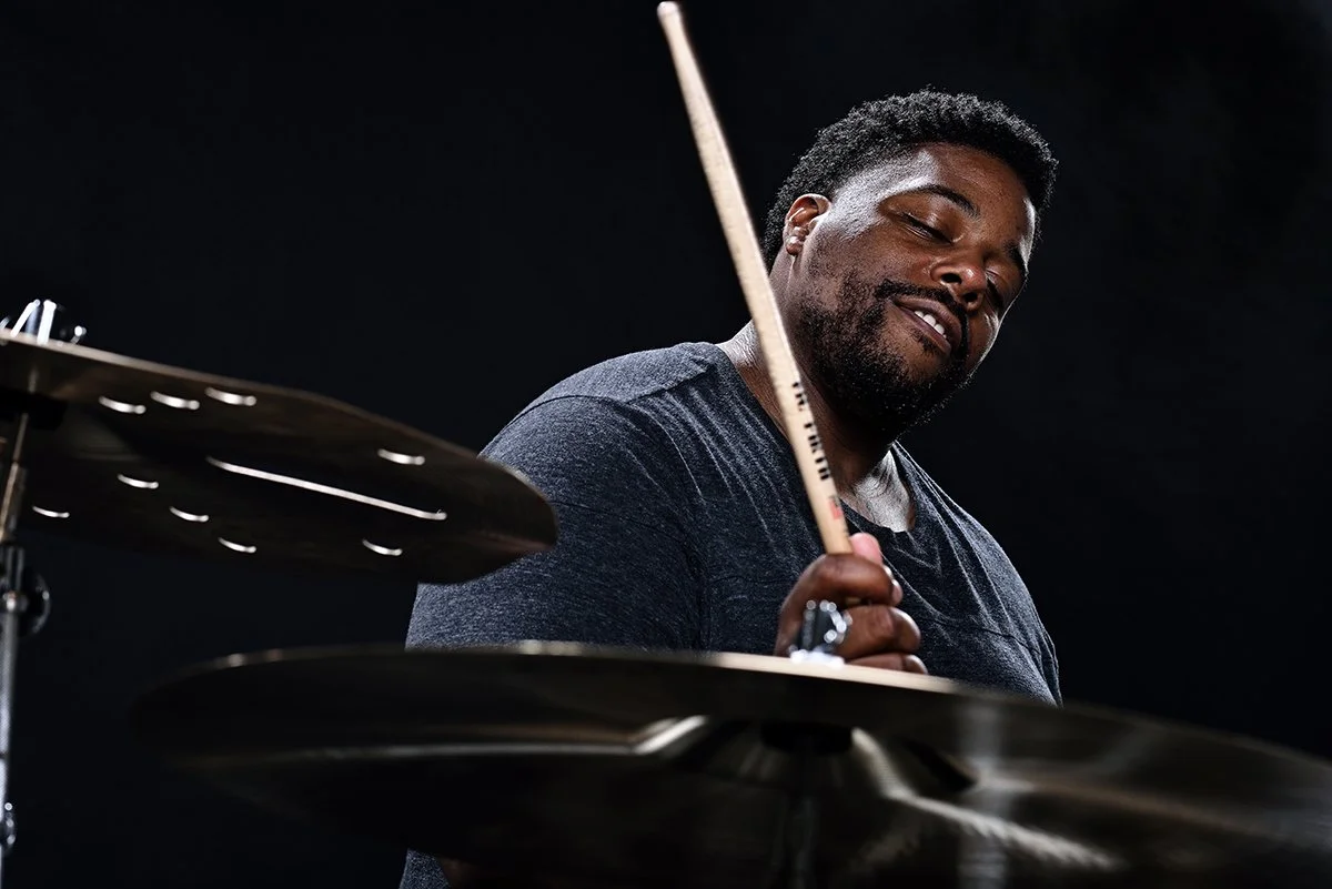 A man playing drums, holding a drumstick, with a focused expression against a dark background.