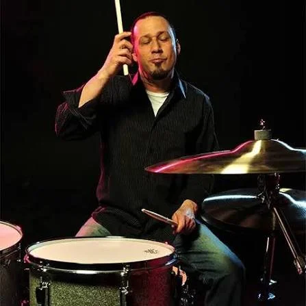 Man playing drums with closed eyes, holding a drumstick to his ear, on a dark stage.