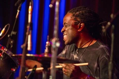 A man with long dreadlocks smiling and playing drums in a music studio.