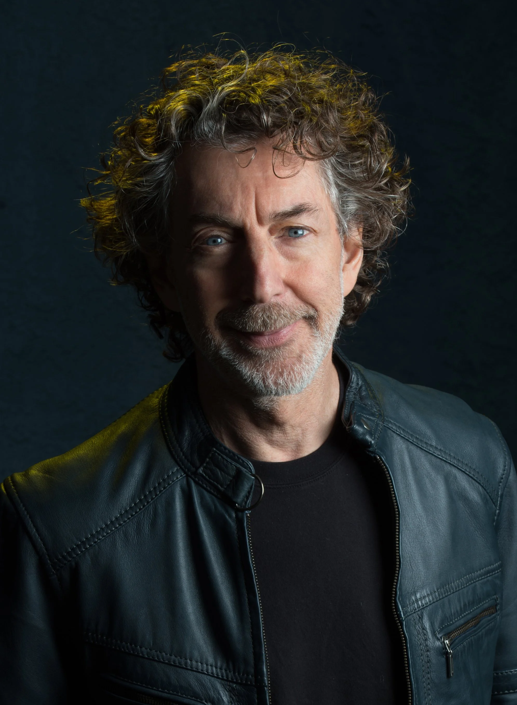 A portrait of a middle-aged man with curly gray hair and blue eyes, wearing a black shirt and a black leather jacket, against a dark background.