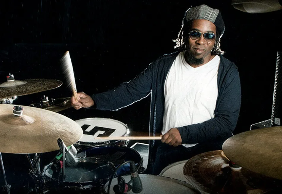 Man wearing glasses, a cap, white shirt, and black hoodie playing drum set with cymbals.