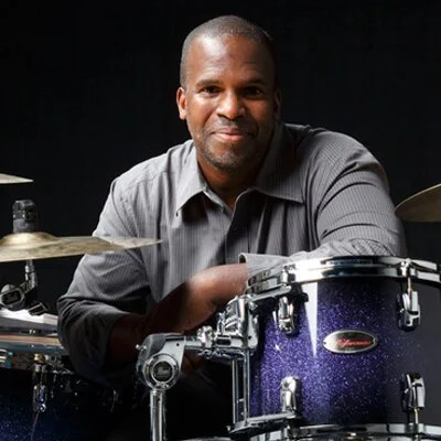 Man sitting behind a drum set, smiling, with a dark background.
