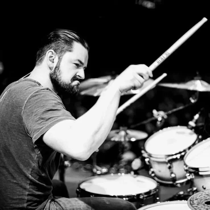 A man with a beard and short hair playing drums in a studio.