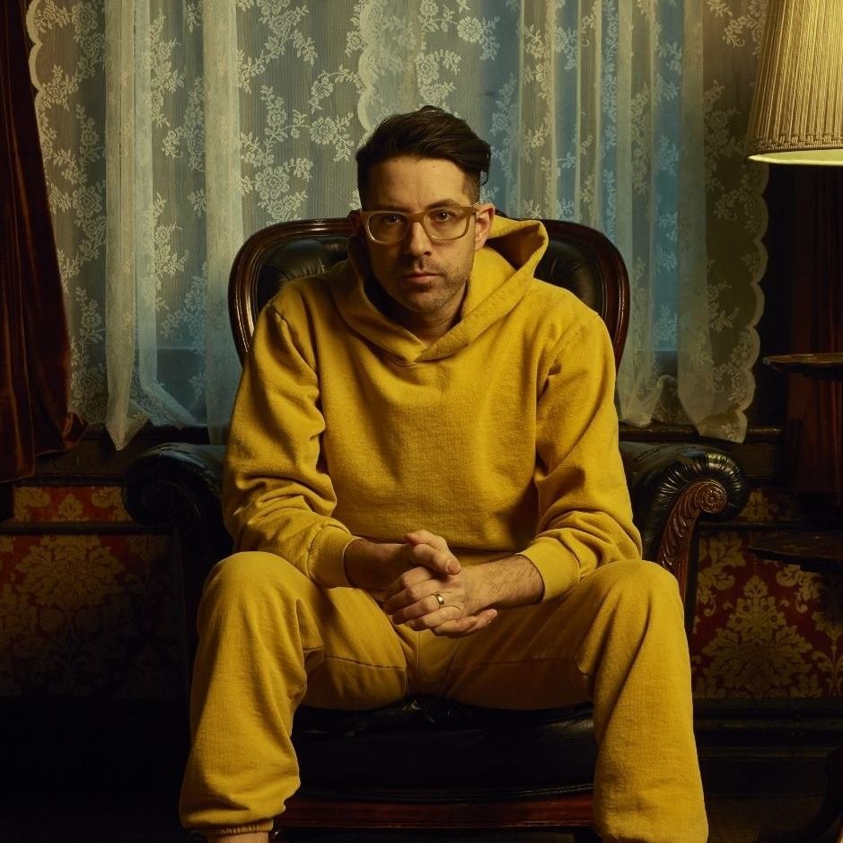 A man wearing glasses and a yellow hoodie sitting in a vintage armchair with ornate wood details, in a room with lace curtains and warm lighting.