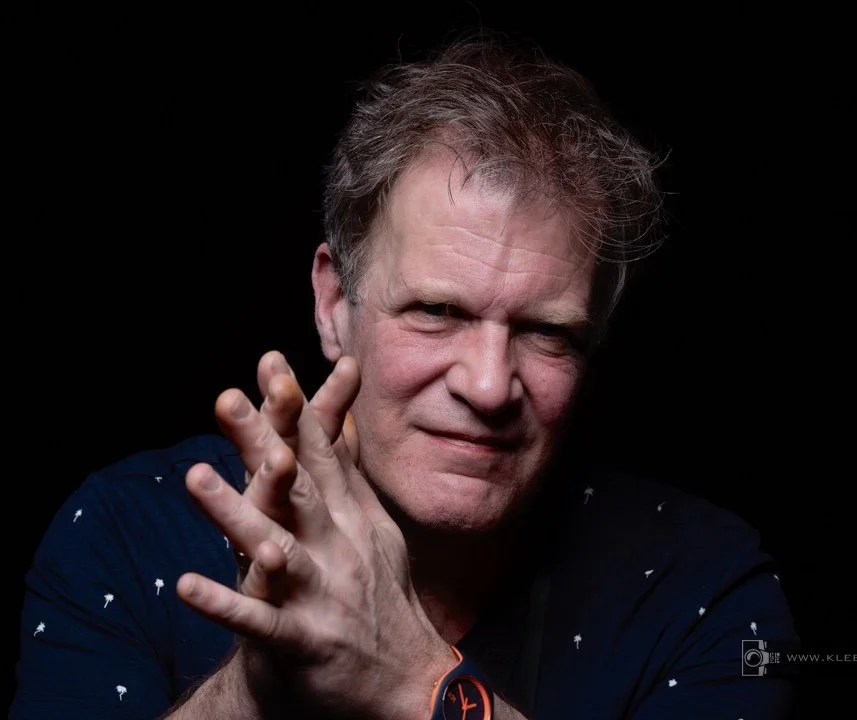 A middle-aged man with disheveled hair and light skin, wearing a dark shirt with small white patterns, is posing against a black background. He has a slight smile and is holding his hand near his face, with his fingers extended.