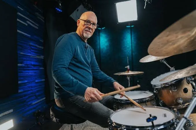 A man sitting at a drum set in a recording studio holding drumsticks, smiling at the camera.