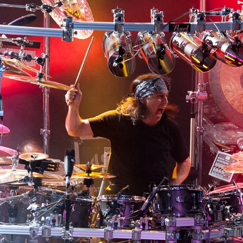 A drummer passionately playing a drum kit on stage, wearing a bandana and black t-shirt, surrounded by stage lights and equipment.