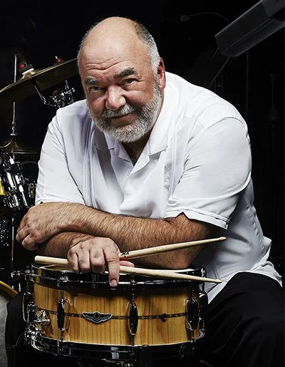 A man with a beard and bald head leaning on a snare drum, holding drumsticks, in a music studio with drums in the background.