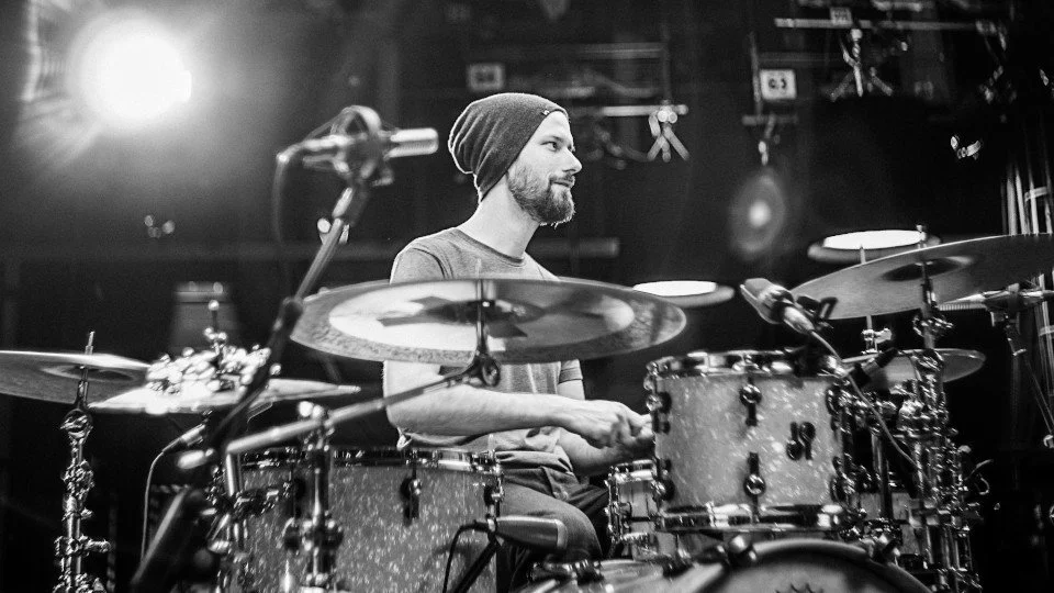 A man with a beard wearing a beanie playing the drums on stage in a black-and-white photo.