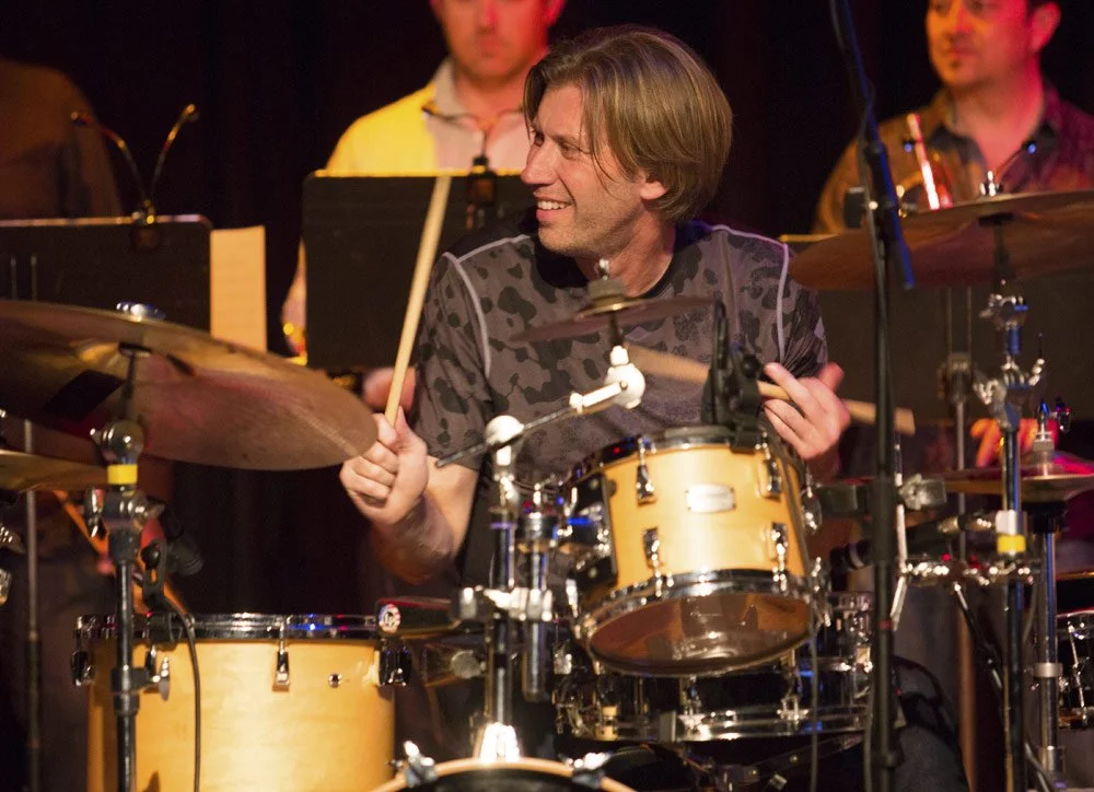 A man with medium-length brown hair smiling while playing a drum set during a band performance, with other band members in the background.