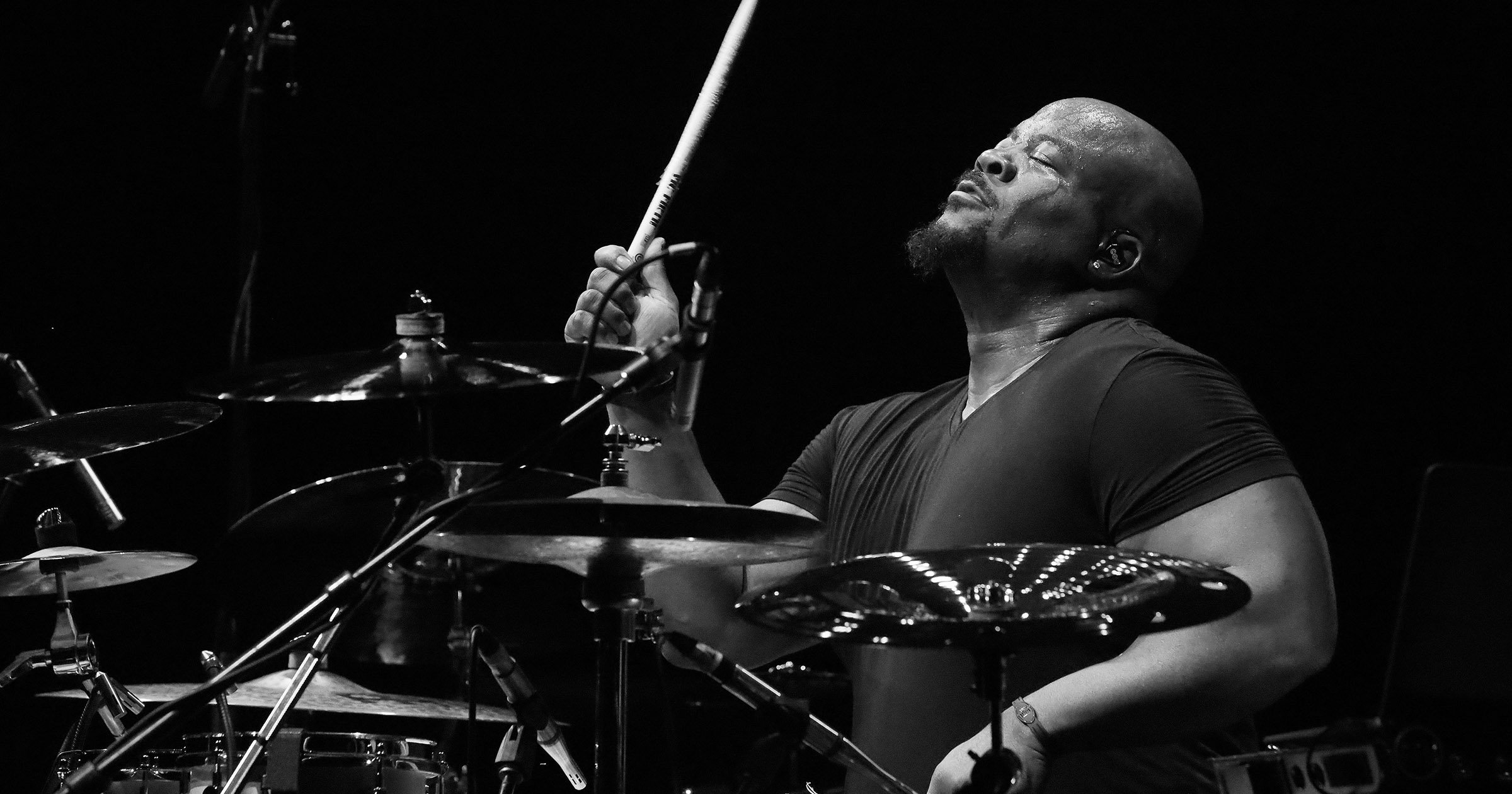 A black and white photo of a male drummer passionately playing the drums, with his eyes closed and holding a drumstick near his face.