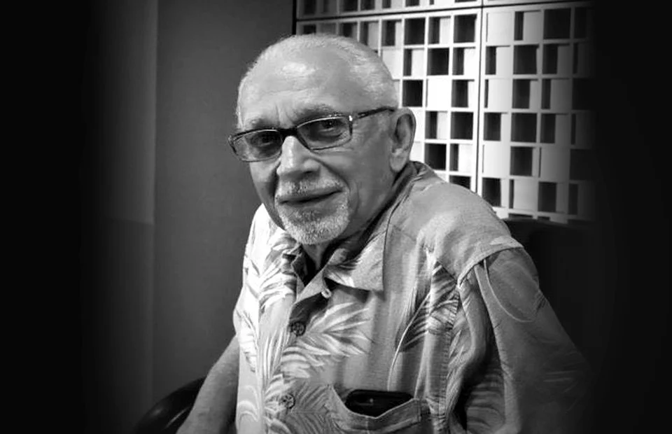Black and white photo of an older man with glasses, short white hair, and a beard, sitting indoors and smiling, wearing a patterned short-sleeve shirt.