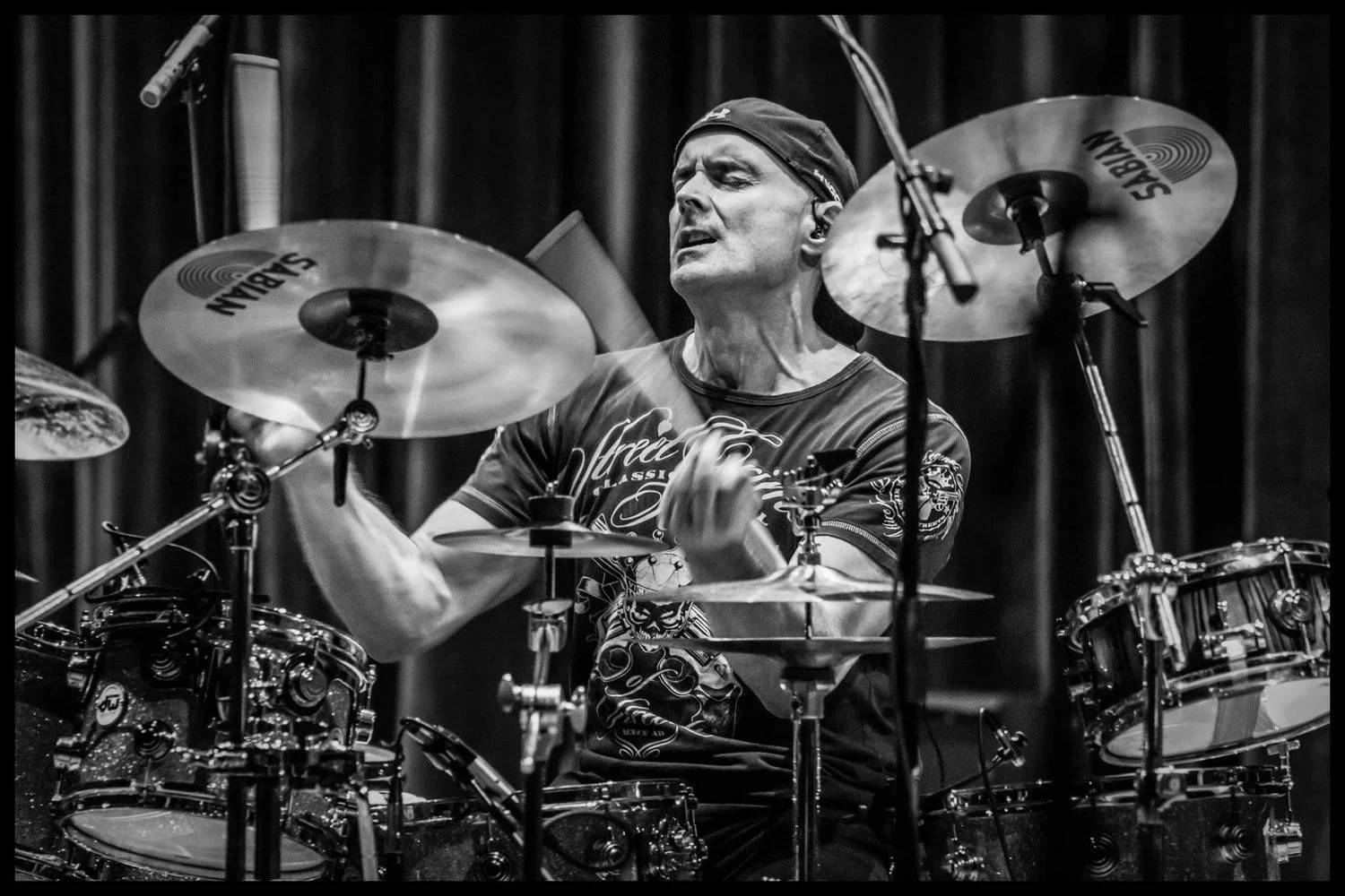 A man playing drums passionately on stage, surrounded by cymbals and drum set, in black and white.