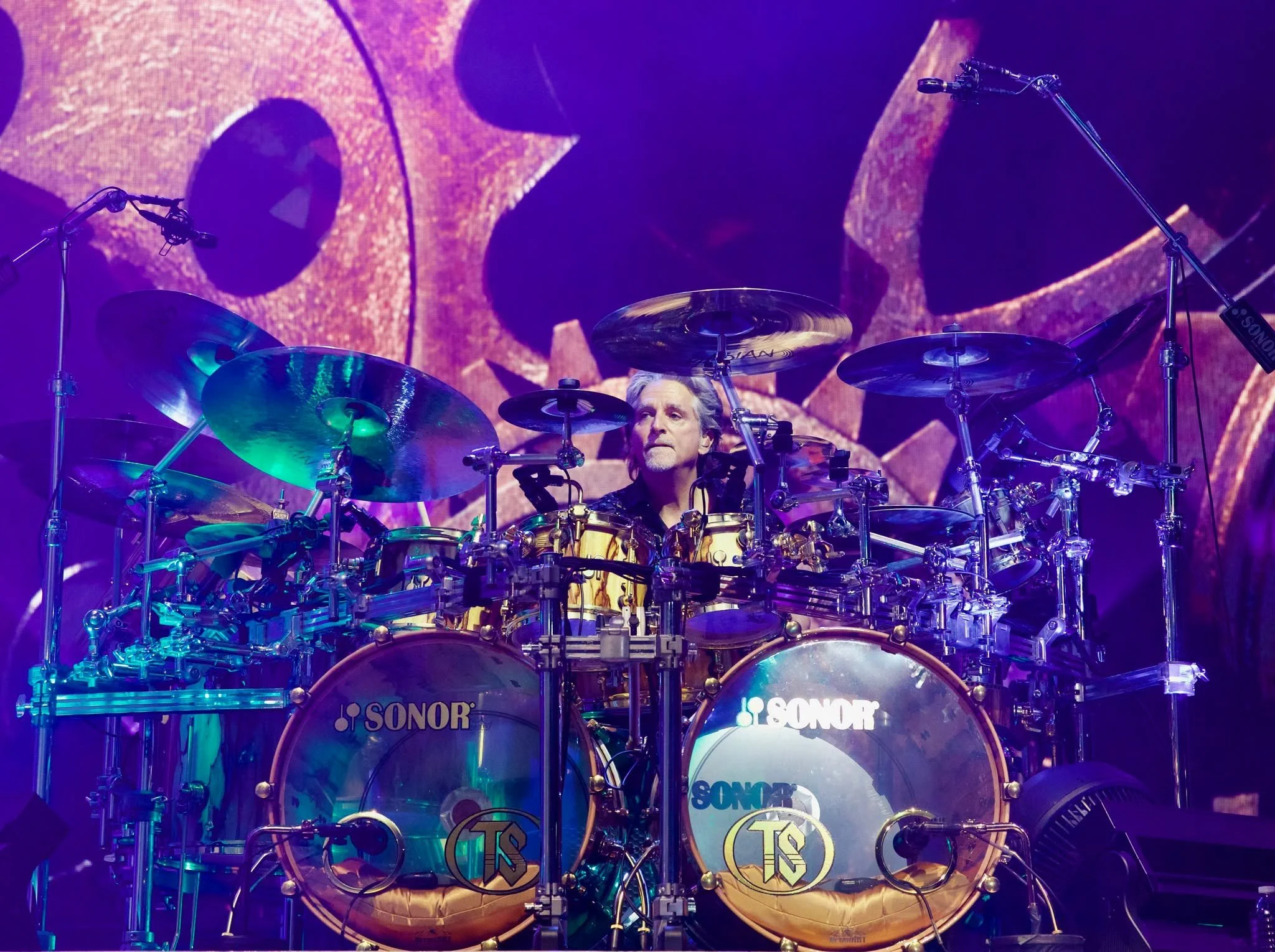 A drummer performing on stage surrounded by a full drum kit with cymbals and drums, with a large screen displaying colorful abstract visuals behind him.