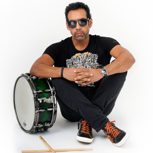 A man sitting on the floor with crossed legs, wearing black sunglasses, a black T-shirt with a graphic design, black jeans, and black sneakers with orange laces. There is a green snare drum and a drumstick next to him.