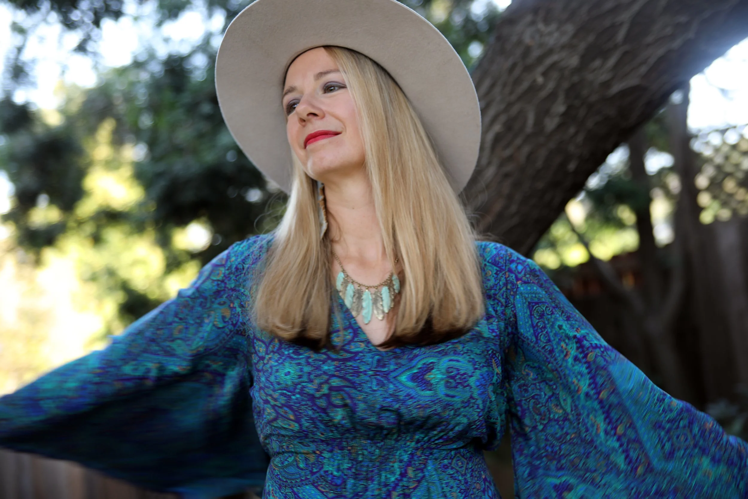 A woman outdoors wearing a large beige hat and a blue patterned dress with a necklace, standing next to a tree with greenery in the background.