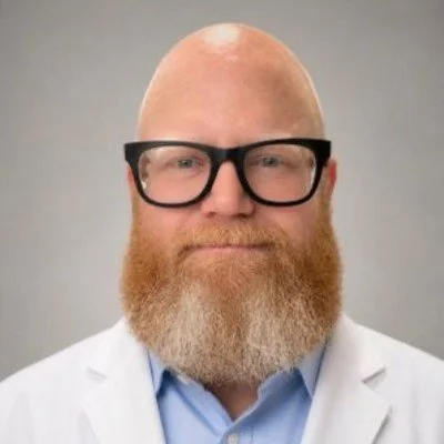 Portrait of a bald man with glasses and a red beard, wearing a white lab coat and a light blue shirt.