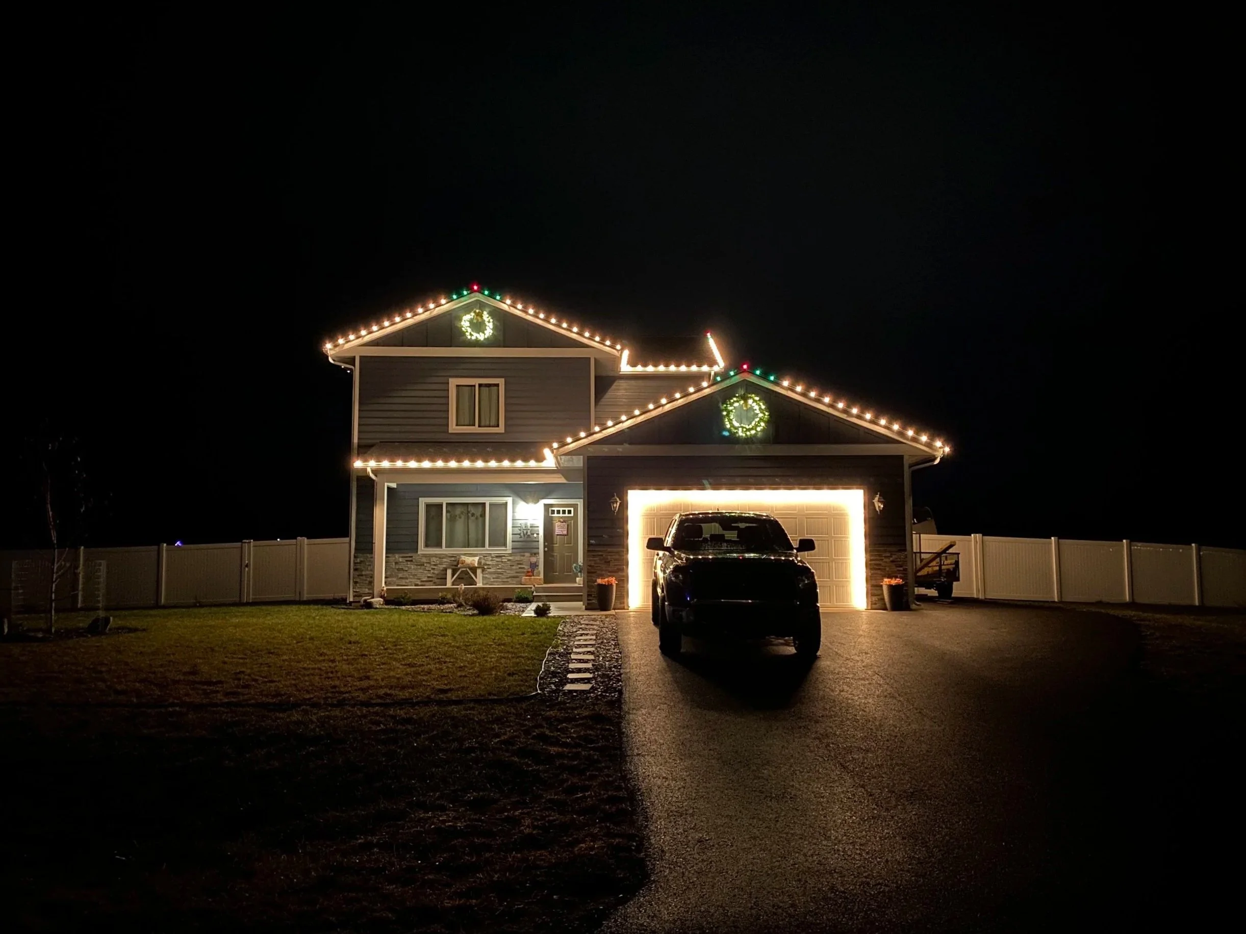 A house decorated with Christmas lights, including a wreath on the second floor window and uplighting on the roof, at night. There is a driveway with a parked car in front of the garage.